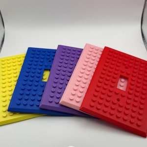 Könnte beinhalten: Ein Set aus fünf rechteckigen Lichtschalterabdeckungen in Gelb, Blau, Lila, Hellrosa und Rot. Jede Abdeckung weist ein erhabenes, kreisförmiges Noppenmuster auf der Oberfläche auf, das an Bausteine erinnert, mit einem zentralen rechteckigen Ausschnitt.