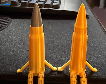 May include: Two 3D-printed novelty items shaped like bullets, each with small arms and legs. One bullet is gold with a grey tip, the other is gold. They are posed as if holding hands.