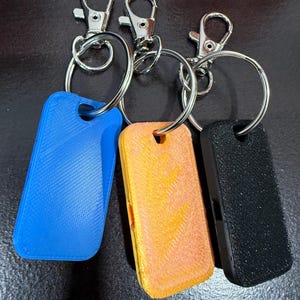 Op de afbeelding: Drie sleutelhangers met rechthoekige, getextureerde labels in blauw, oranje en zwart. Elk label heeft een klein gaatje aan de bovenkant en is bevestigd aan een zilveren metalen ring en een karabijnhaak.