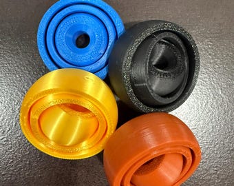 May include: Four 3D-printed objects in blue, black, yellow, and orange. Each object has a circular shape with concentric rings and a central opening. The objects are arranged on a dark surface.
