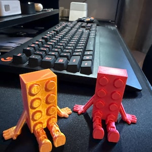 May include: Two 3D-printed figures resembling building blocks, one orange and one red, each with arms and legs. The orange figure is on the left, and the red figure is on the right. The figures are on a black surface.