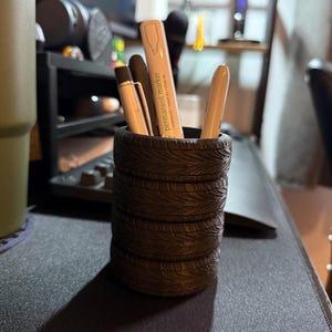 Puede incluir: Un organizador de escritorio negro con forma de neumáticos apilados, que contiene bolígrafos y un rotulador permanente. El rotulador es blanco y beige con las palabras "permanent marker" impresas. El organizador está sobre una superficie oscura.