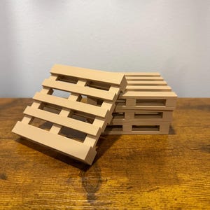 May include: A stack of miniature, light brown wooden pallet coasters. One coaster is angled, showing the slatted design. The coasters are on a wooden surface.