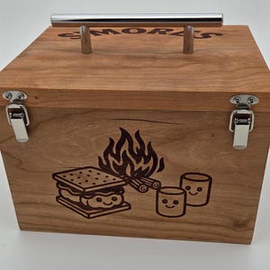 May include: A rectangular wooden box with a silver metal handle on top and two silver metal latches on the front. The lid features the engraved text S'MORES. The front panel displays an engraved graphic of a campfire, a s'more, and two marshmallows.