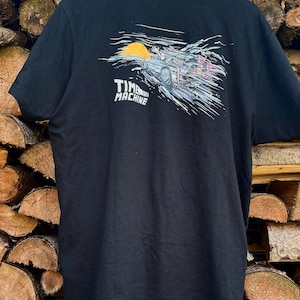 Puede incluir: Camiseta negra con un gráfico de un DeLorean, una máquina del tiempo, que acelera a través de una puesta de sol. El texto "TIME MACHINE" está impreso debajo del coche.