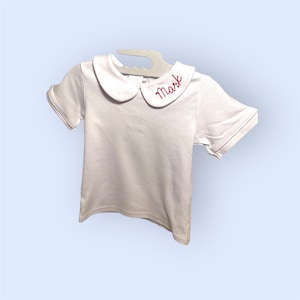 May include: White short-sleeved shirt with a Peter Pan collar. The collar has the name "Mark" embroidered in red thread. The shirt is hanging on a white hanger against a light blue background. The shirt is made of a soft material.