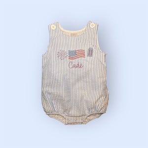 Könnte beinhalten: Blau-weiß gestreifter Seersucker-Strampler mit gestickter amerikanischer Flagge, Feuerwerk und dem roten Namen "Cade". Der Strampler hat Knopfverschlüsse an den Schultern und einen Druckknopfverschluss am unteren Ende.