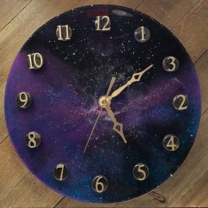 May include: Round wall clock with a galaxy design. The clock face features a deep blue and purple background with gold-colored numerals and hands. The clock is approximately 12 inches in diameter.