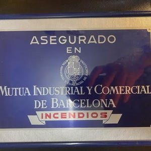 Puede incluir: Un letrero rectangular azul y plateado con marco azul. El texto dice ASEGURADO EN MUTUA INDUSTRIAL Y COMERCIAL DE BARCELONA INCENDIOS, e incluye un emblema circular central con un escudo de armas y el nombre de la empresa en letras blancas.