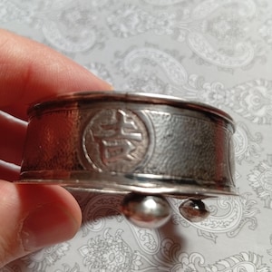 Puede incluir: Un recipiente circular de metal plateado con una superficie texturizada y un emblema circular en relieve que presenta un carácter chino. El recipiente tiene un borde liso y se apoya sobre pequeñas patas redondeadas en forma de bola.