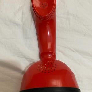 Könnte beinhalten: Ein leuchtend roter, glänzender Telefonhörer aus Kunststoff mit geschwungenem, ergonomischem Design. Der obere Teil verfügt über ein Lautsprechergitter mit fünf kleinen Löchern, während die Basis ein gewölbtes, perforiertes Gehäuse ist.