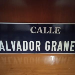 Puede incluir: Una placa de calle rectangular de metal azul oscuro con letras blancas que dicen CALLE SALVADOR GRANES. En el lado izquierdo, hay un pequeño escudo con un árbol y una corona sobre un fondo azul con un borde blanco.