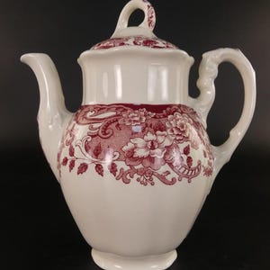 Op de afbeelding: Een witte keramische theepot met een donkerrood bloemenpatroon rondom de buik en het deksel. De theepot heeft een gebogen handvat, een spitse tuit en een bijpassend deksel met een lusvormige knop, geplaatst tegen een donkere achtergrond.