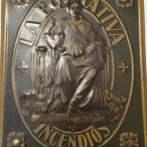 Puede incluir: Una placa metálica rectangular con acabado verde oscuro y relieve en color bronce. El centro presenta una figura sentada sosteniendo una balanza, rodeada por el texto LA EQUITATIVA, FUNDACION ROSILLO e INCENDIOS, con detalles florales en las esquinas.