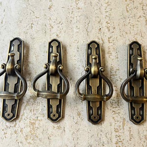 May include: Four antique brass door pulls with a decorative design. Each pull features a rectangular backplate with a central handle and a curved, arched handle. The brass has a warm, aged patina.