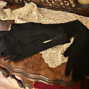 May include: A pair of black gloves with a long, elegant design. The gloves are made of a soft material and feature a buttoned cuff detail. They are laid out on a wooden surface with a cream-colored lace.