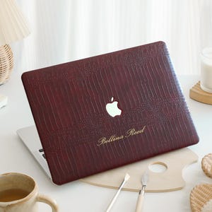 Op de afbeelding: Een donkerbordeauxkleurige laptophoes met een gestructureerd krokodillenleerpatroon. De hoes is voorzien van een uitsparing voor het Apple-logo en de goudkleurige tekst Bellina Reed die onderaan in het midden op het oppervlak is gedrukt.