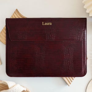 May include: A rectangular dark burgundy laptop sleeve with a textured crocodile skin pattern and a flap closure. The name Laura is printed in gold lettering on the center of the flap. The sleeve has a slim profile and a structured design.