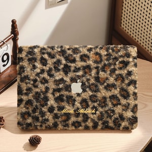May include: A laptop case with a leopard print design in shades of brown and black. The Apple logo is visible, along with the name "Mark Aldrich" in a script font. The case is on a wooden table.