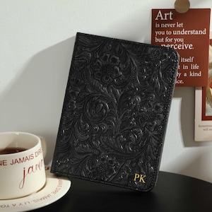 May include: Black leather journal with embossed floral pattern and gold initials "PK". A white teacup with the words "NE JAMAIS DIRE jamais" is on a saucer. A decorative art print is in the background.