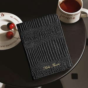 May include: A black rectangular passport holder with a textured crocodile skin pattern and the name Yilia Kevin embossed in gold script on the bottom right corner. The item is positioned next to a white ceramic mug and a small white plate with cherries.