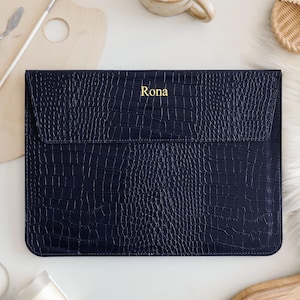 May include: A dark navy blue laptop sleeve with a textured crocodile skin pattern and a flap closure. The name Rona is printed in gold lettering on the center of the flap. The sleeve has a rectangular shape with rounded corners and stitched edges.