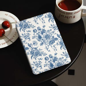 Könnte beinhalten: Eine rechteckige, cremefarbene Hülle mit blauem Blumenmuster. Das Design zeigt detaillierte blaue Blumen und Blätter. Die Hülle liegt auf einer schwarzen Oberfläche, in der Nähe einer weißen Tasse mit Text und einem Teller mit Kirschen.