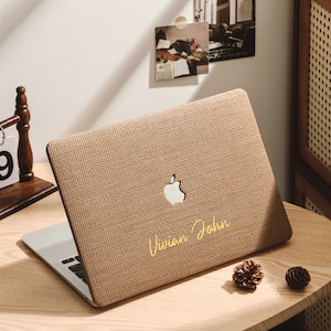 Może przedstawiać: Beżowy laptop z teksturowaną obudową, logo Apple i imieniem "Vivian John" złotym pismem. Leży na drewnianym biurku obok drewnianego kalendarza z napisem "DEC 39".