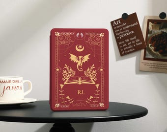 Puede incluir: Una funda roja con un diseño de dragón y libro dorado, con las iniciales "RJ." Una taza blanca con las palabras "JAMAIS DIRE jamas" está a la izquierda. Un letrero marrón con la palabra "Art" está a la derecha.