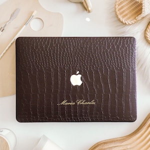 May include: A dark brown laptop cover with a textured crocodile skin pattern. The cover features a central cutout for the Apple logo and the name Maria Charles engraved in gold lettering on the lower portion of the rectangular surface.