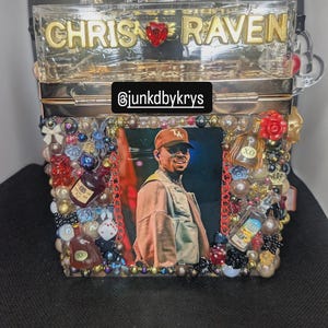 May include: A clear, rectangular box purse with the names "CHRIS" and "RAVEN" in gold lettering. The purse is decorated with a photo, beads, and miniature liquor bottles. The Instagram handle "@junkdbykrys" is also visible.