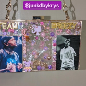 May include: Clear acrylic clutch with gold-tone chain strap. The clutch features images of a singer, decorative beads, and the words "TEAM DEUCES BREEZY". The interior is filled with pink and purple embellishments.
