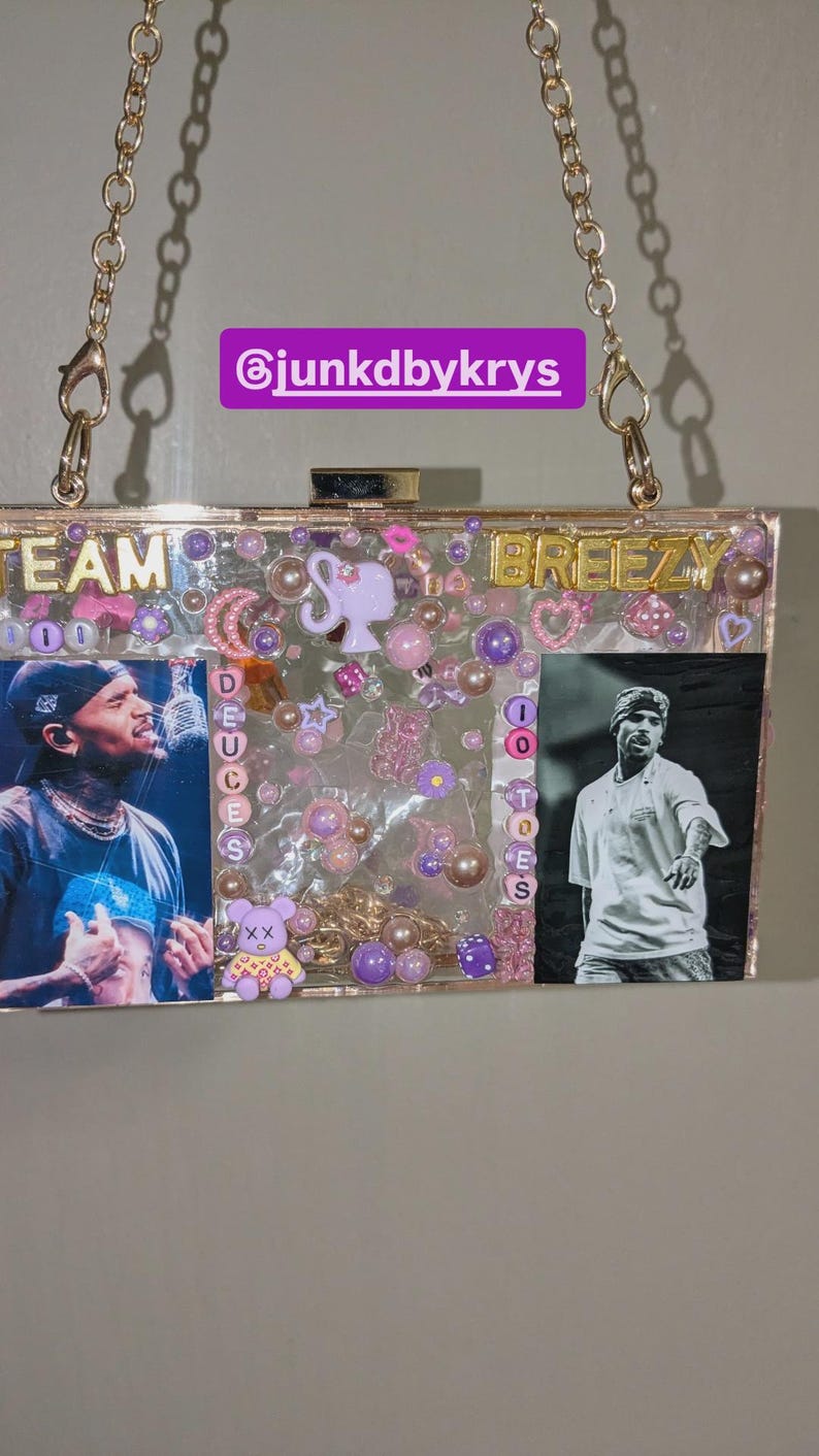May include: Clear acrylic clutch with gold-tone chain strap. The clutch features images of a singer, decorative beads, and the words "TEAM DEUCES BREEZY". The interior is filled with pink and purple embellishments.