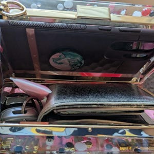 May include: A clear, rectangular clutch with gold-colored trim, holding a dark phone case with a pop socket, a wallet, keys, and a pink object. The clutch has a decorative pattern on the bottom.