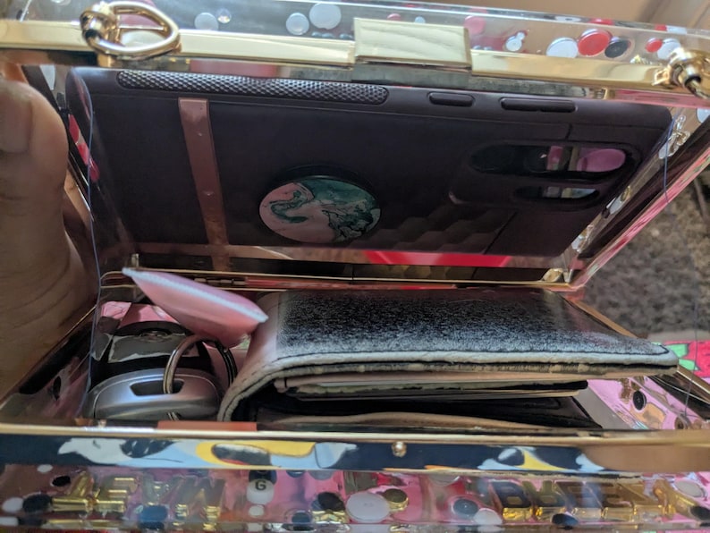 May include: A clear, rectangular clutch with gold-colored trim, holding a dark phone case with a pop socket, a wallet, keys, and a pink object. The clutch has a decorative pattern on the bottom.