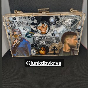 May include: A clear acrylic clutch decorated with images of celebrities, bottle caps, and rhinestones. The clutch features the text "CHANEL", "PRADA", and "GOOD KISSER". The Instagram handle "@junkdbykrys" is also visible.