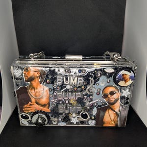 May include: Clear acrylic clutch with silver hardware. The clutch features images of two men, the word "BUMP" in silver letters, and decorative embellishments. The clutch has a silver chain strap.