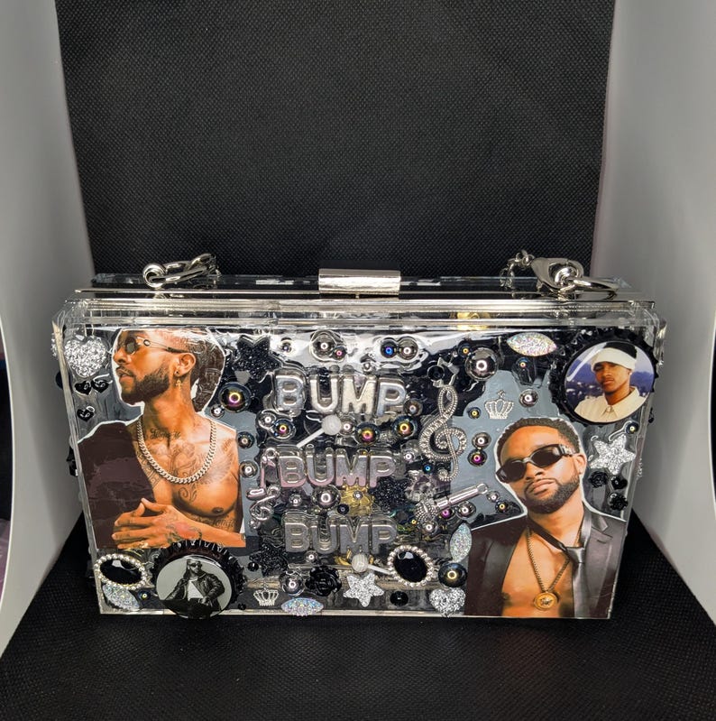 May include: Clear acrylic clutch with silver hardware. The clutch features images of two men, the word "BUMP" in silver letters, and decorative embellishments. The clutch has a silver chain strap.