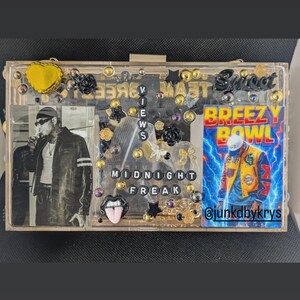 May include: A clear acrylic clutch decorated with gold, black, and clear embellishments. The clutch features a black and white photo, the words "Breezy Bowl" and "Midnight Freak", and the Instagram handle @junkdbykrys.