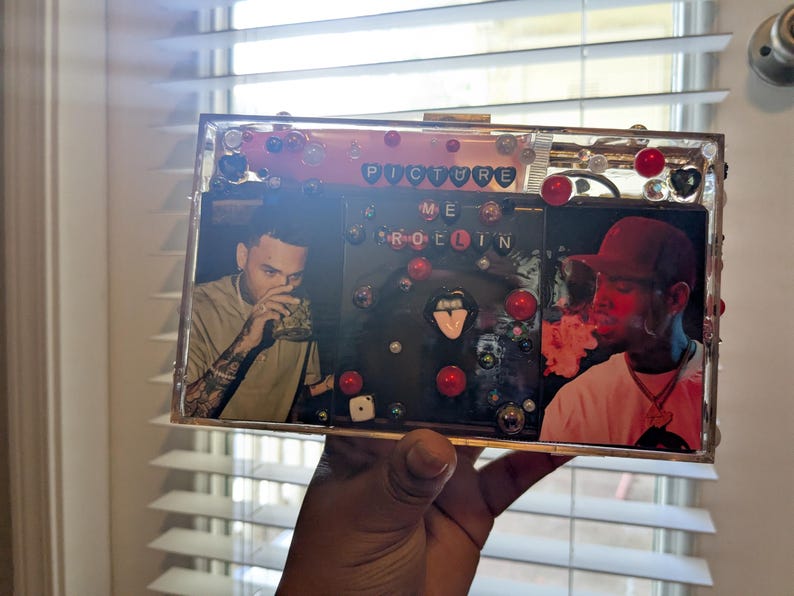 May include: Clear rectangular clutch with images of two people, red and black accents, and the words "PICTURE ME ROLLIN". The clutch is decorated with red beads and other embellishments.