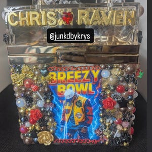 May include: A clear acrylic box clutch with gold lettering that reads "CHRIS RAVEN". The front of the clutch features a "BREEZY BOWL" graphic with a person in a yellow jacket. The clutch is embellished with beads and red roses.