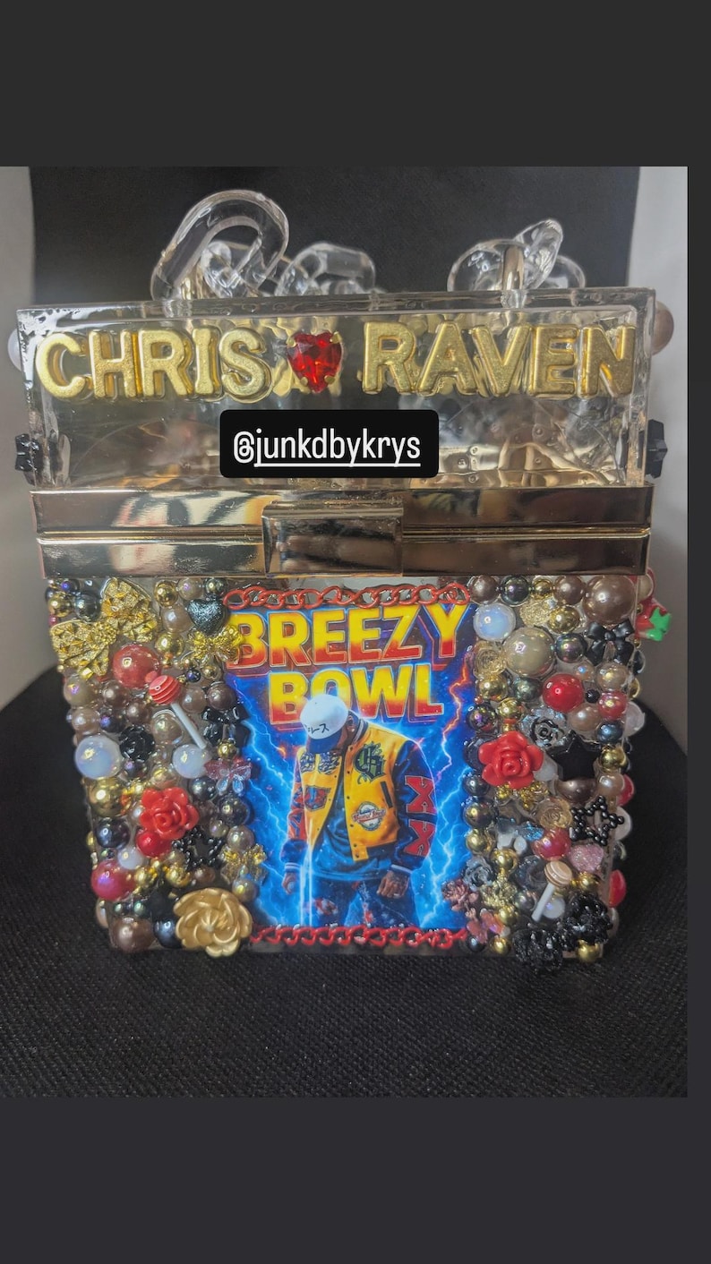 May include: A clear acrylic box clutch with gold lettering that reads "CHRIS RAVEN". The front of the clutch features a "BREEZY BOWL" graphic with a person in a yellow jacket. The clutch is embellished with beads and red roses.