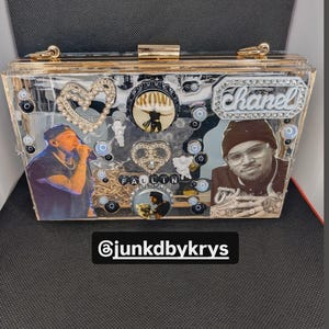 May include: A clear acrylic clutch with gold-colored trim and embellishments. The clutch features images of people, decorative charms, and the text "Chanel". The text "FALLIN" is also visible, along with the Instagram handle "@junkdbykrys".