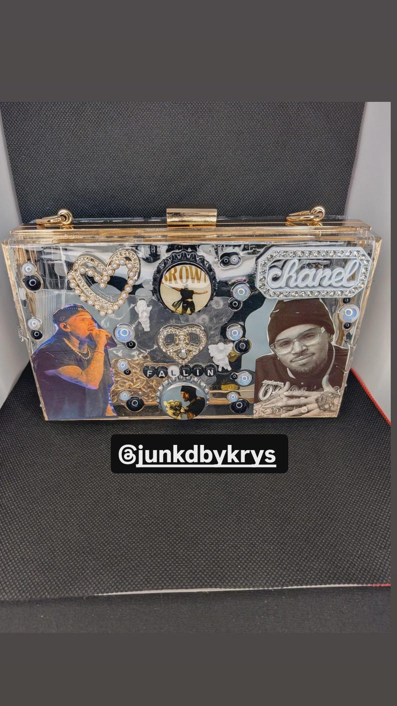 May include: A clear acrylic clutch with gold-colored trim and embellishments. The clutch features images of people, decorative charms, and the text "Chanel". The text "FALLIN" is also visible, along with the Instagram handle "@junkdbykrys".