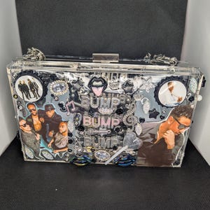 May include: Clear rectangular clutch with silver hardware and chain strap. The clutch is decorated with images of a musical group, the word "BUMP" in multiple places, and various embellishments including lips, stars, and musical notes.