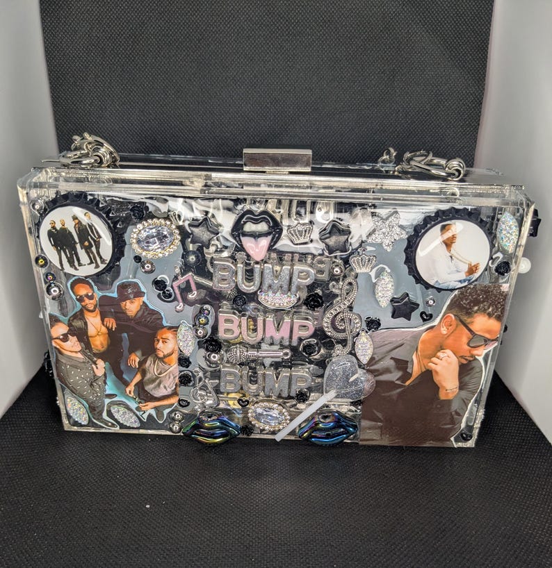 May include: Clear rectangular clutch with silver hardware and chain strap. The clutch is decorated with images of a musical group, the word "BUMP" in multiple places, and various embellishments including lips, stars, and musical notes.