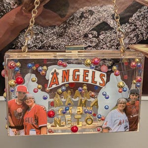 May include: Clear acrylic clutch with gold chain strap. The clutch features the text "ANGELS" and "MAMA NETO" with photos of people wearing Angels baseball apparel. The clutch is decorated with colorful beads.