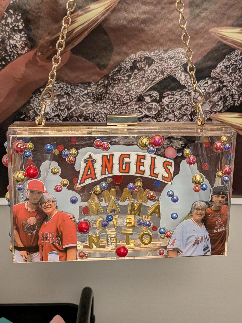 May include: Clear acrylic clutch with gold chain strap. The clutch features the text "ANGELS" and "MAMA NETO" with photos of people wearing Angels baseball apparel. The clutch is decorated with colorful beads.