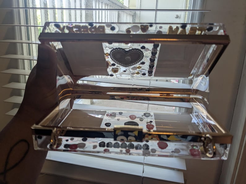 May include: A clear, rectangular box with gold trim and a hinged lid. The lid and sides are decorated with colorful, irregular shapes. The interior features a heart-shaped design and a small latch. The word "DREAMS" is visible on the top.