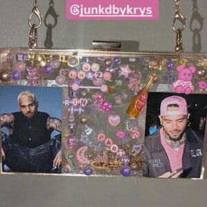 May include: Clear acrylic clutch with gold chain strap, filled with pink, purple, and clear embellishments. Features photos of a person, along with the words "RUN IT CRAZY PARTY". Includes a small champagne bottle charm.